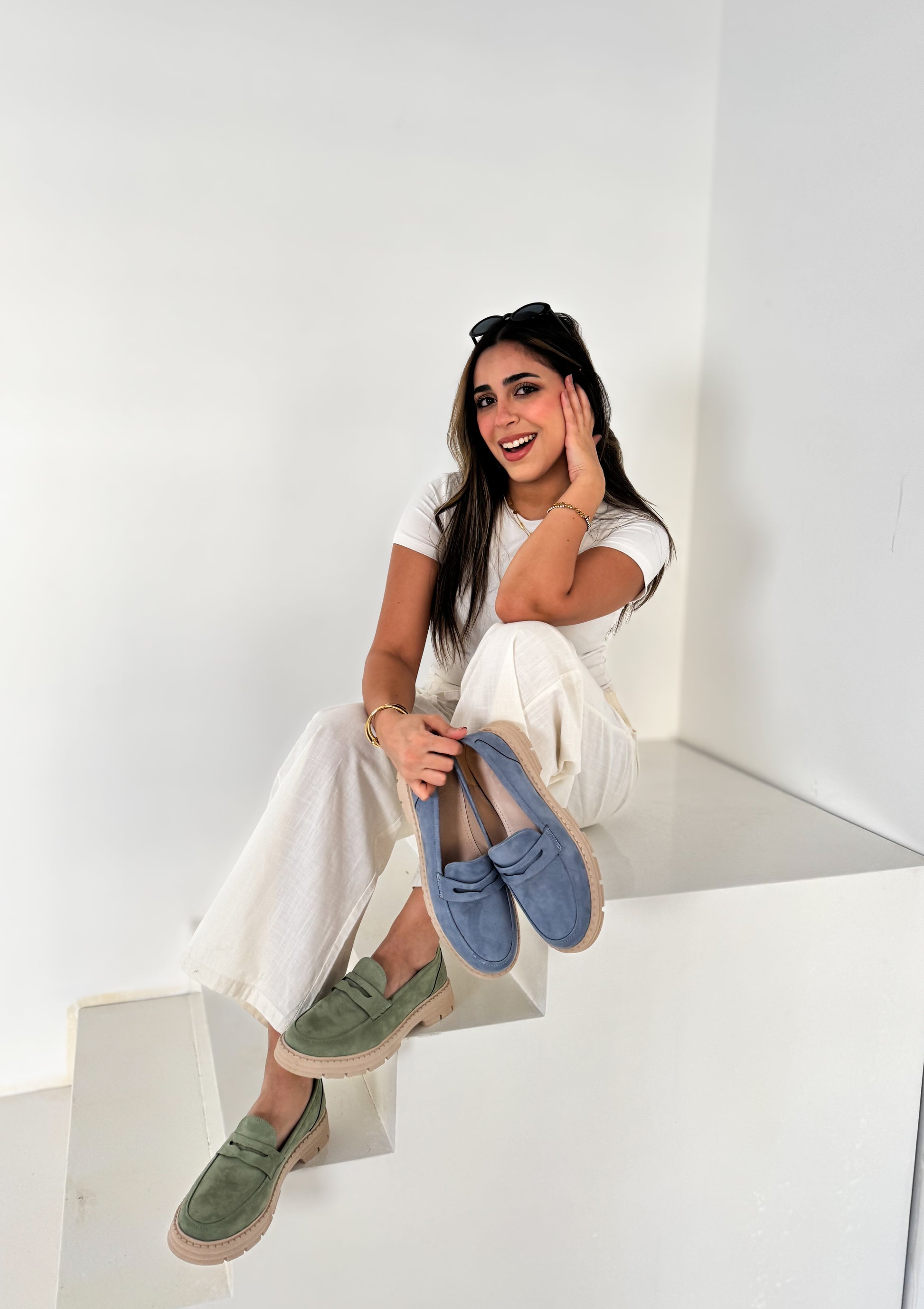 Woman sitting on a white cube wearing green and blue  shoes.