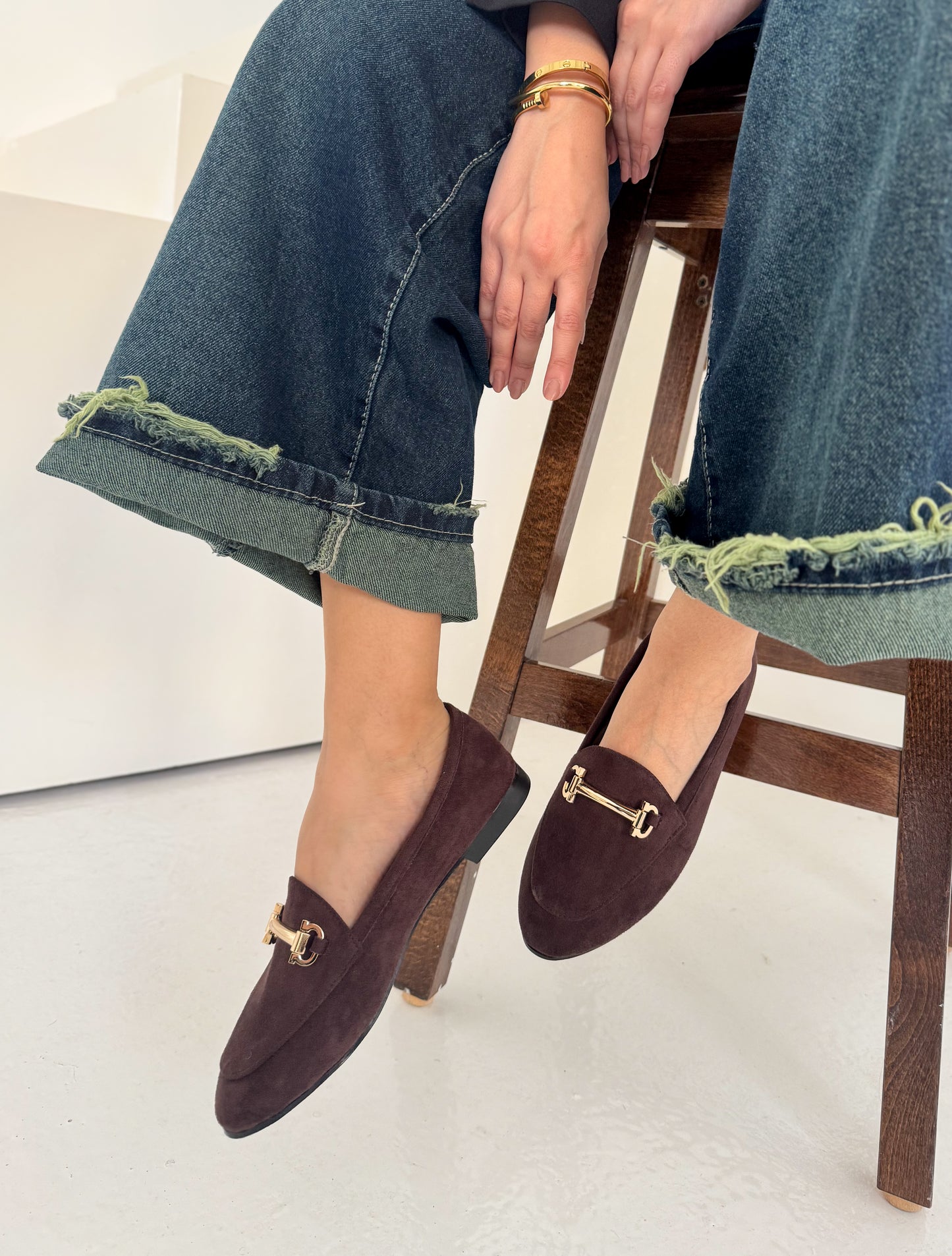 Brown loafers with gold buckles worn by a woman sitting on a wooden stool.