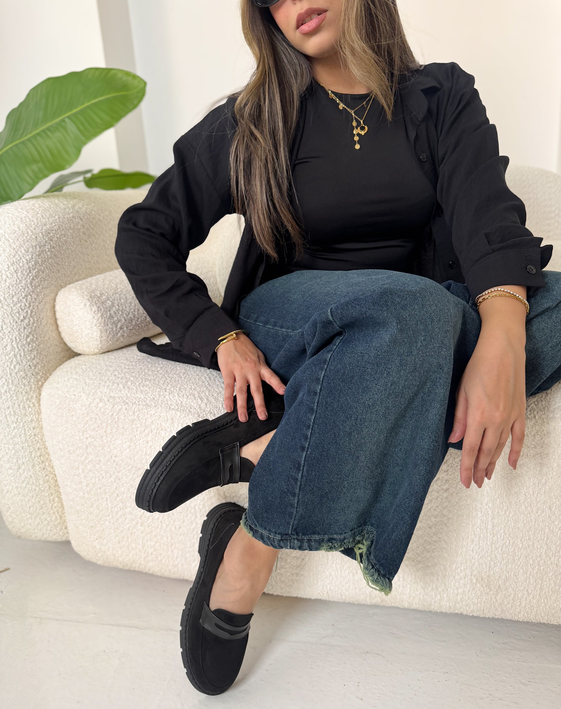 woman sitting on a white couch wearing black shoes, black jacket, and blue jeans.