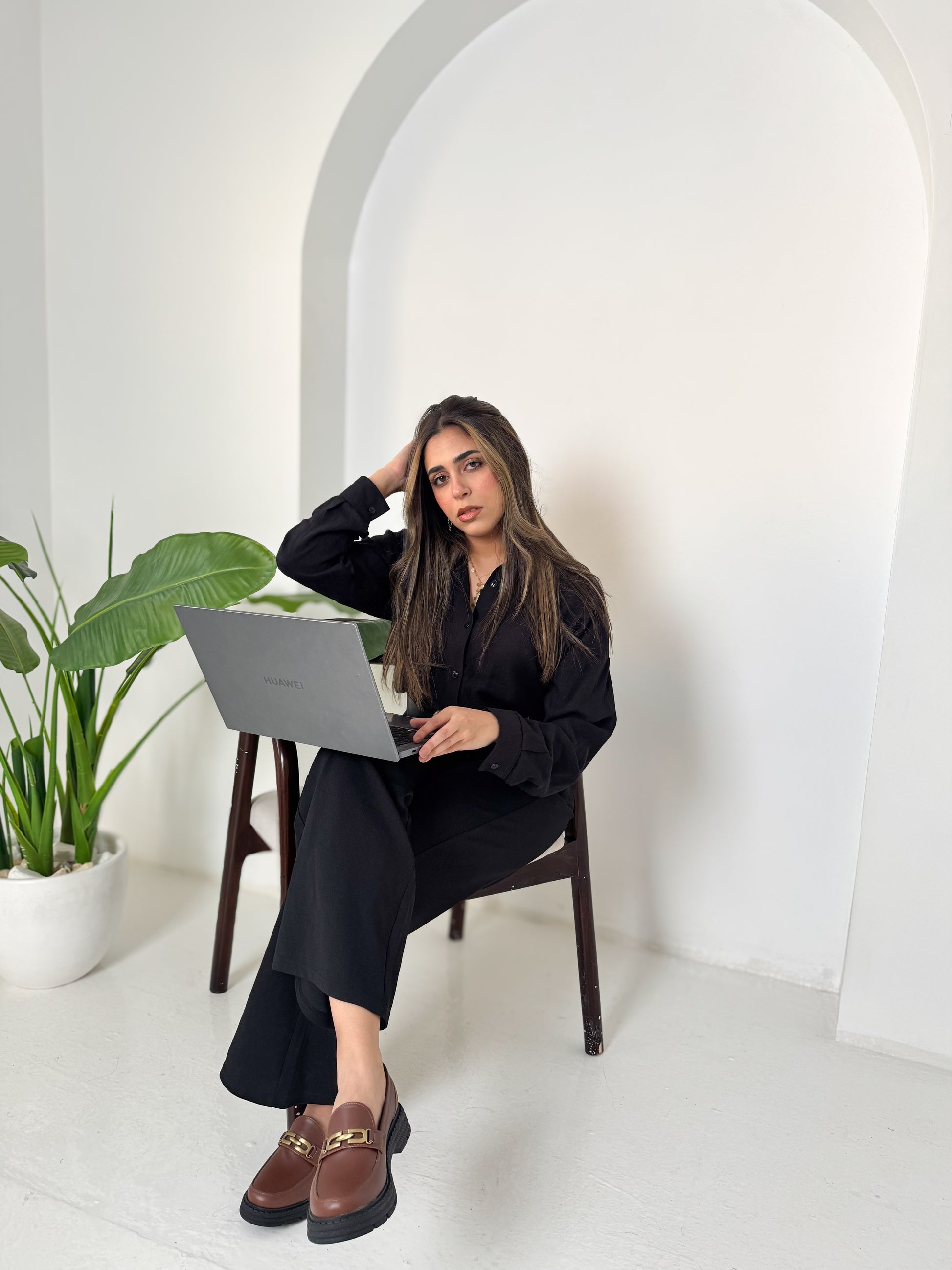 Woman sitting on a chair with a laptop in a minimalistic room with white walls and a plant.