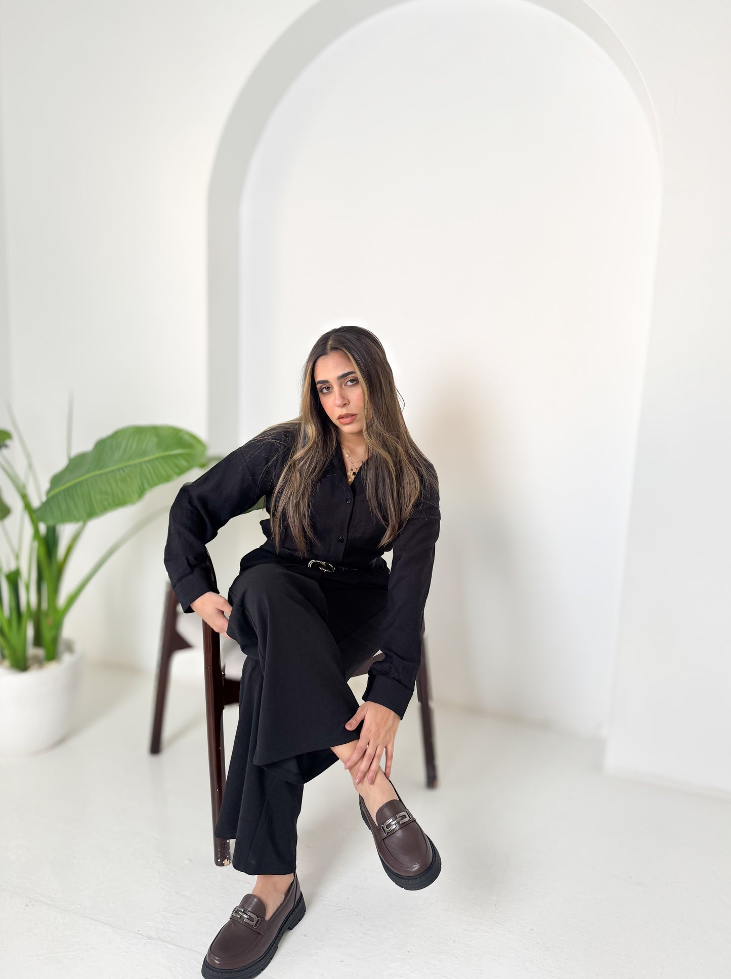Woman in a black outfit sitting on a chair in a minimalistic setting with a plant in the background.