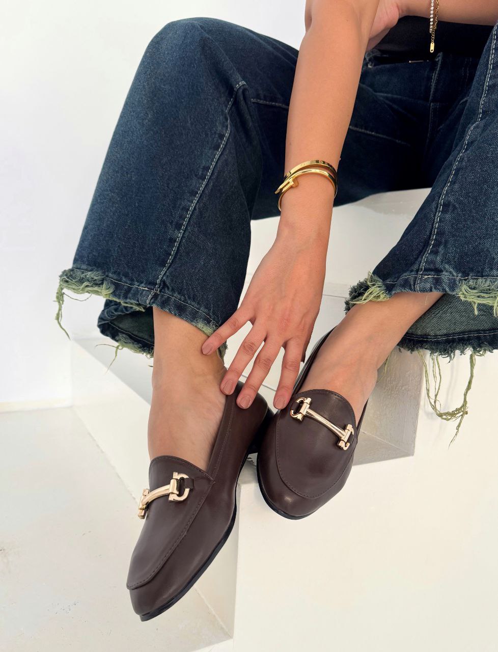 Brown loafers with gold accents worn by a woman sitting on a white surface.
